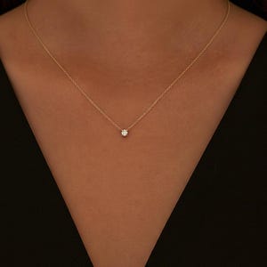 May include: A delicate gold necklace featuring a small, sparkling diamond pendant. The necklace is a simple design, with a thin chain and a single, round-cut diamond. The necklace is worn against a dark background.