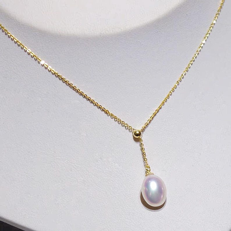 May include: A delicate gold necklace featuring a single, lustrous, oval-shaped pearl pendant. The pearl has a soft, iridescent sheen. The chain is a fine, linked design, and the necklace is displayed against a white background.