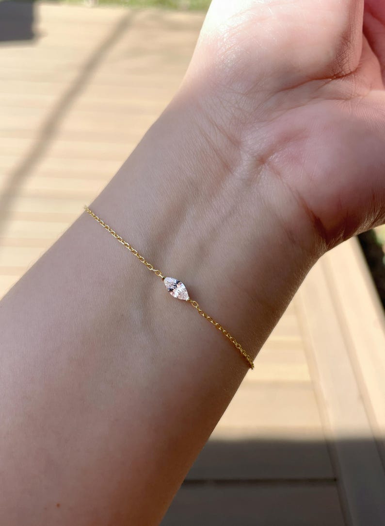 Pu&ograve; includere: Un delicato braccialetto d'oro con una gemma taglio marquise. Il braccialetto &egrave; una catena sottile con una gemma centrale, allungata e trasparente. Il gioiello &egrave; esposto su un polso, mostrando l'elegante design del braccialetto.