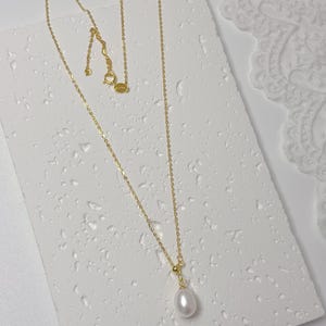 May include: A delicate gold necklace with a single, teardrop-shaped white pearl pendant. The necklace chain is fine and the clasp is visible. The jewelry is displayed on a textured white surface.