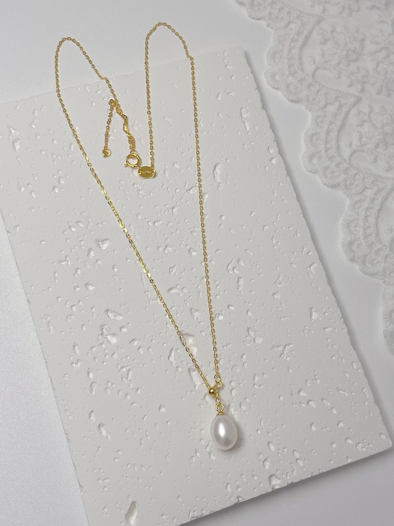 May include: A delicate gold necklace with a single, teardrop-shaped white pearl pendant. The necklace chain is fine and the clasp is visible. The jewelry is displayed on a textured white surface.