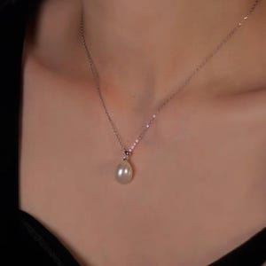 May include: A delicate silver necklace featuring a single, luminous, teardrop-shaped pearl pendant. The pearl hangs gracefully from a fine chain, creating a classic and elegant look. The necklace is shown against a dark background.