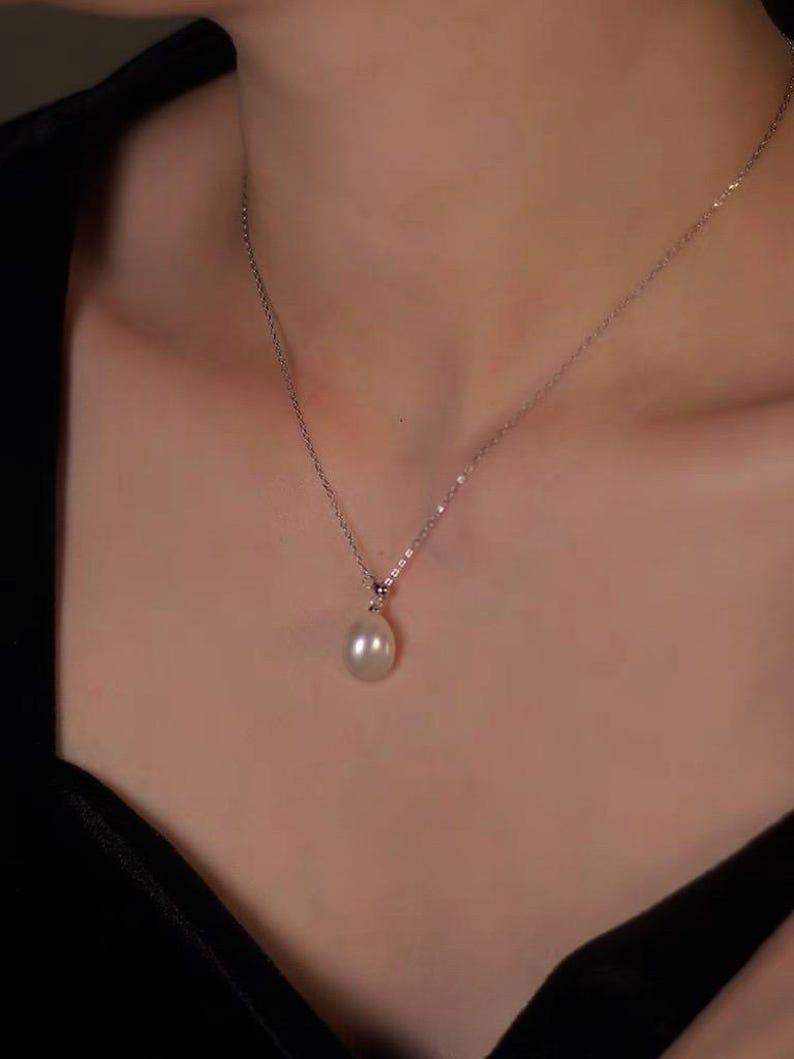 May include: A delicate silver necklace featuring a single, luminous, teardrop-shaped pearl pendant. The pearl hangs gracefully from a fine chain, creating a classic and elegant look. The necklace is shown against a dark background.