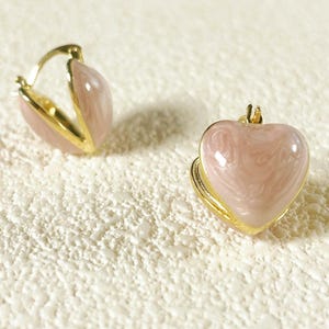 May include: Gold-tone heart-shaped earrings with a pink marbled enamel finish. One earring is open, revealing the gold interior. The earrings are set against a textured, off-white surface. Jewelry.