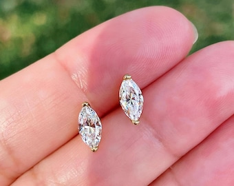 May include: A pair of stud earrings featuring marquise-cut clear gemstones set in gold-toned metal. Each earring has a pointed oval shape with a faceted surface that reflects light, secured by a small gold-toned prong at the top and bottom ends.