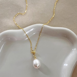 May include: A delicate gold-colored necklace featuring a single, teardrop-shaped white pearl pendant. The chain has a subtle, textured design. The necklace is displayed on a white, curved surface, creating a minimalist aesthetic.