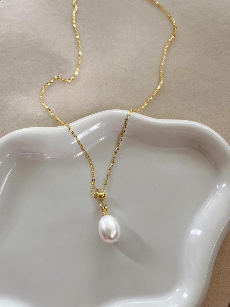May include: A delicate gold-colored necklace featuring a single, teardrop-shaped white pearl pendant. The chain has a subtle, textured design. The necklace is displayed on a white, curved surface, creating a minimalist aesthetic.