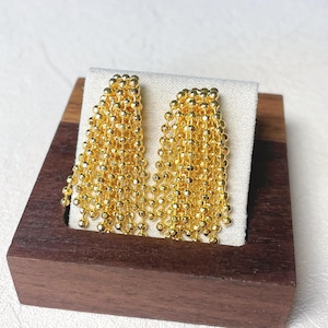 May include: A pair of gold-colored dangle earrings. Each earring features multiple strands of small, round beads cascading downwards. The earrings are displayed on a white fabric, set within a dark wooden box.