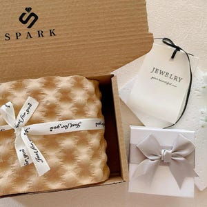 Pu&ograve; includere: Una confezione regalo con la scritta "SPARK" e il logo, contenente un articolo avvolto legato con un nastro con la scritta "Just for you". Una piccola scatola bianca con un fiocco grigio e un'etichetta con la scritta "JEWELRY".