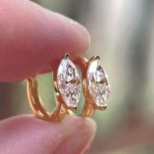 May include: A pair of gold hoop earrings, each featuring a single marquise-cut clear gemstone set in a polished gold frame. The earrings are designed as small hoops with a hinged closure, showcasing a bright, reflective finish on the metal.