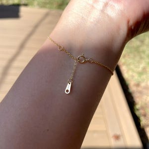 Pu&ograve; includere: Un delicato braccialetto d'oro con una piccola chiusura e una catena regolabile &egrave; mostrato. Il braccialetto &egrave; esposto al polso di una persona, con la luce del sole che si riflette sull'oro. Il braccialetto sembra essere un gioiello raffinato.
