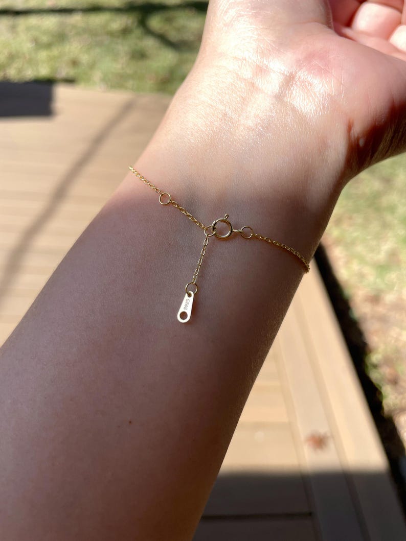 Pu&ograve; includere: Un delicato braccialetto d'oro con una piccola chiusura e una catena regolabile &egrave; mostrato. Il braccialetto &egrave; esposto al polso di una persona, con la luce del sole che si riflette sull'oro. Il braccialetto sembra essere un gioiello raffinato.