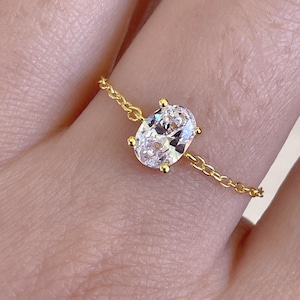 May include: A gold ring featuring an oval-cut, clear gemstone. The ring's band is a delicate gold chain. The gemstone is held in place by four prongs. The ring is on a finger.