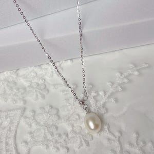 May include: A delicate silver necklace featuring a single, teardrop-shaped pearl pendant. The pearl is a creamy white color and hangs from a fine chain. The necklace is displayed against a white, textured background.