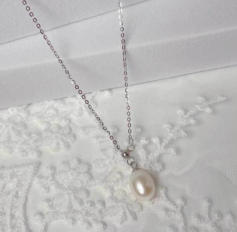 May include: A delicate silver necklace featuring a single, teardrop-shaped pearl pendant. The pearl is a creamy white color and hangs from a fine chain. The necklace is displayed against a white, textured background.