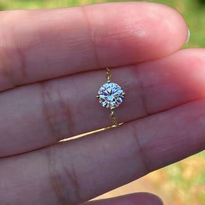 May include: A delicate gold necklace featuring a single, round, clear gemstone. The gemstone is faceted, catching the light and creating a sparkling effect. The necklace is held by a person's hand.