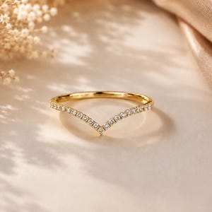 May include: A gold V-shaped ring featuring a row of small, clear, round-cut stones set along the band. The ring has a polished metallic finish and a pointed chevron design, creating a symmetrical appearance with multiple sparkling accents.