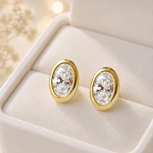 May include: A pair of oval-shaped stud earrings featuring clear, faceted stones set in polished yellow gold bezels. The earrings are displayed side-by-side, showcasing the smooth, rounded metal edges surrounding each brilliant-cut center stone.