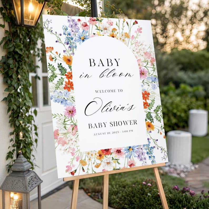 Puede incluir: Un letrero de bienvenida para baby shower blanco sobre un caballete de madera, adornado con un borde floral de flores silvestres coloridas. El letrero dice "Baby in Bloom" y da la bienvenida a los invitados al baby shower de Olivia, con la fecha y hora.