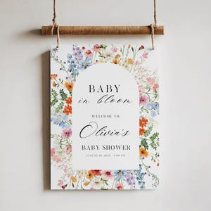 Puede incluir: Un letrero de baby shower con el texto "BABY in bloom" y "Welcome to Olivia's BABY SHOWER August 10, 2025 - 1:00 PM". El letrero est&aacute; adornado con flores coloridas y est&aacute; colgado de una varilla de madera con cuerda.