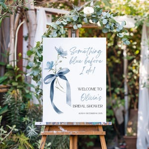 May include: A white sign on a wooden easel with the text "Something blue before I do!" and "Welcome to Olivia's Bridal Shower". The sign features blue floral illustrations and a blue ribbon. The date is Sunday, December 10, 2025.