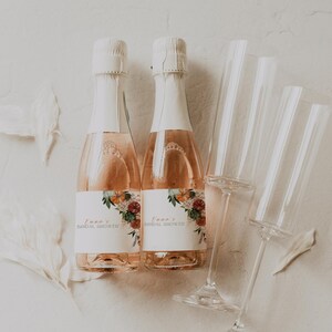 May include: Two champagne bottles with white labels and floral designs, labeled "Emma's Bridal Shower." The bottles are next to several clear champagne flutes. The champagne has a pink hue, and the bottles are sealed with white foil.
