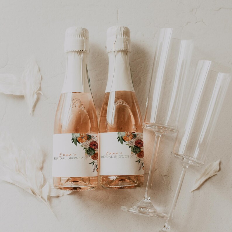 May include: Two champagne bottles with white labels and floral designs, labeled "Emma's Bridal Shower." The bottles are next to several clear champagne flutes. The champagne has a pink hue, and the bottles are sealed with white foil.