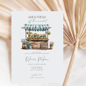 Puede incluir: Una tarjeta de invitación azul claro con el texto "SHE'S FRESH off the market" sobre una ilustración en acuarela de un puesto de flores. La tarjeta incluye detalles para una despedida de soltera en honor a Olivia Wilson, con fecha, hora e información de RSVP.