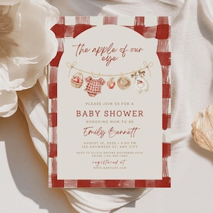 Pu&ograve; includere: Un invito rettangolare per baby shower con bordo a quadri rossi e bianchi. Il centro mostra un pannello color crema ad arco con il testo The apple of our eye, illustrato con un cesto di mele, una tutina, una mela, un cappello e un coniglio. La carta include i dettagli dell'evento per Emily Bennett il 10 agosto 2025 alle 15:00, con informazioni su RSVP e registro in rosso e marrone scuro.