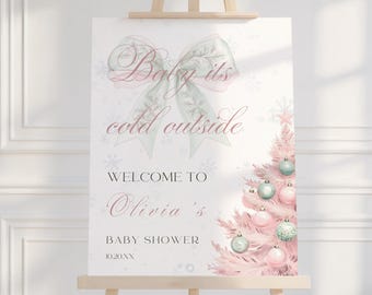 May include: A baby shower sign on a wooden easel. The sign features the text "Baby its cold outside" above "Welcome to Olivia's Baby Shower" and a date. A pink Christmas tree with ornaments is on the right.