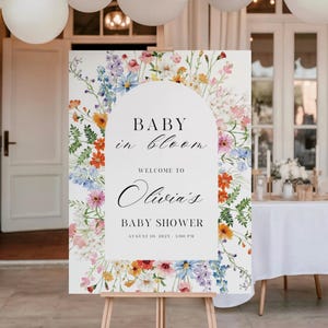 Puede incluir: Un letrero blanco sobre un caballete de madera con el texto "BABY in bloom" y "Welcome to Olivia's BABY SHOWER August 10, 2023 - 3:00 PM". El letrero est&aacute; decorado con arreglos florales coloridos en tonos rosa, naranja, azul y amarillo.