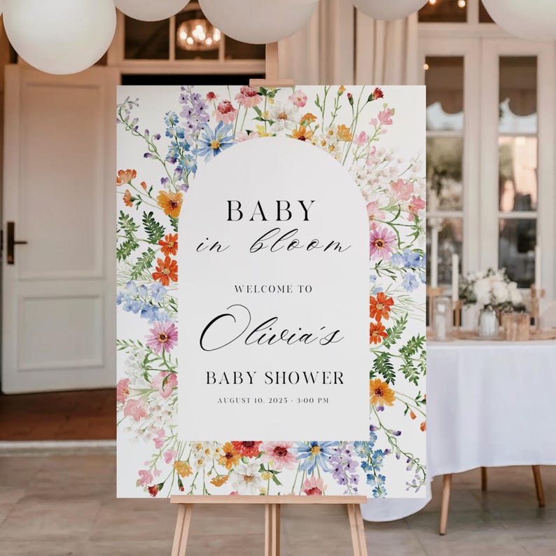Puede incluir: Un letrero blanco sobre un caballete de madera con el texto "BABY in bloom" y "Welcome to Olivia's BABY SHOWER August 10, 2023 - 3:00 PM". El letrero est&aacute; decorado con arreglos florales coloridos en tonos rosa, naranja, azul y amarillo.