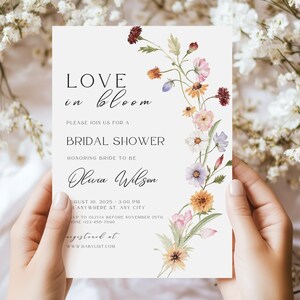 May include: A bridal shower invitation with the words "Love in Bloom" and floral accents. The invitation includes details such as the date, time, and location, along with the bride's name, Olivia Wilson. The invitation is held by a person.