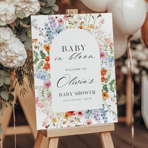 Puede incluir: Un letrero blanco con borde floral y el texto "BABY in bloom" y "Bienvenida a la fiesta de bienvenida de Olivia". El letrero se muestra en un caballete de madera. El borde floral incluye flores rosas, naranjas, azules y moradas.