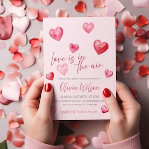 May include: A pink bridal shower invitation with watercolor hearts and the phrase "love is in the air." The invitation includes the name Olivia Wilson, date, time, and RSVP information. Registered at Target & Amazon.