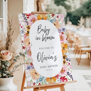 May include: A floral baby shower welcome sign on an easel. The sign features a white oval with the text "Baby in Bloom" and "Welcome to Olivia's Baby Shower 09.20.25". The background is a watercolor floral design with pink, orange, and blue flowers.