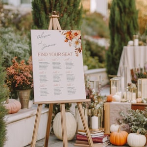 May include: A wedding seating chart on a wooden easel. The sign reads "Olivia and James" and "Find Your Seat." Floral accents in orange and white decorate the top right corner. Pumpkins and candles are in the background.
