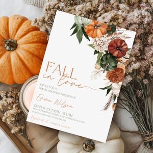 May include: A white invitation card with the text "Fall in Love" and details for a bridal shower. The card is decorated with watercolor pumpkins and floral elements in shades of orange, green, and brown. A pumpkin and candle are in the background.
