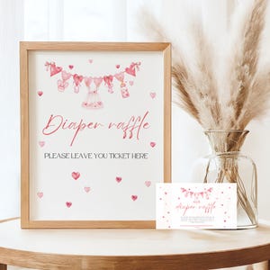May include: A framed sign with the words "Diaper raffle" in pink script, decorated with baby-themed illustrations and hearts. The sign also includes the text "PLEASE LEAVE YOU TICKET HERE". A smaller matching card is next to the frame.