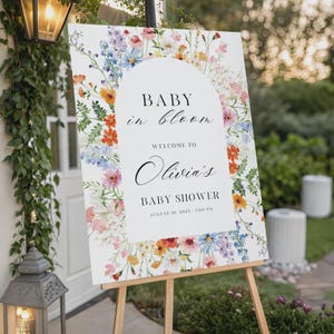 以下が含まれることがあります： 木製のイーゼルに立てられた白い看板。テキスト「Baby in Bloom」と「Welcome to Olivia's Baby Shower」が書かれています。カラフルな花柄のボーダーで飾られています。