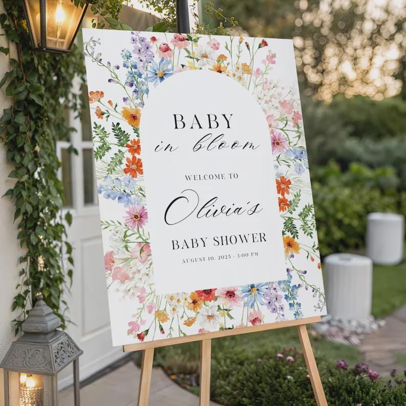 以下が含まれることがあります： 木製のイーゼルに立てられた白い看板。テキスト「Baby in Bloom」と「Welcome to Olivia's Baby Shower」が書かれています。カラフルな花柄のボーダーで飾られています。