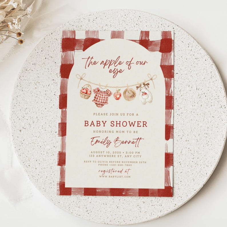 Pu&ograve; includere: Un biglietto d'invito rettangolare per baby shower con un bordo a quadri rossi e bianchi. Il centro presenta uno sfondo color crema ad arco con il testo 'The apple of our eye' sopra un'illustrazione di un filo da bucato con articoli.