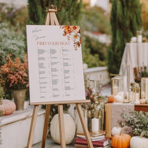 May include: A wooden easel displays a seating chart for Olivia and James, with the text "Find Your Seat." The chart features a floral design and a list of names. Pumpkins and candles decorate the outdoor setting.