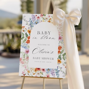 Puede incluir: Un letrero de baby shower con detalles florales y el texto "Baby in Bloom". El letrero blanco tiene un dise&ntilde;o arqueado y presenta flores coloridas. Se adjunta un gran lazo color crema.