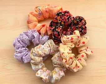 May include: Five colorful scrunchies with floral patterns. The scrunchies are made of soft fabric and are perfect for holding hair in place.