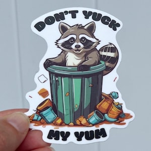 Sticker being held by hand. Cute raccoon in trash can with the words "Don't Yuck My Yum".