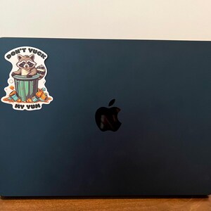 Sticker on black Apple laptop. Sticker pictures cute raccoon in trash can with the words "Don't Yuck My Yum".