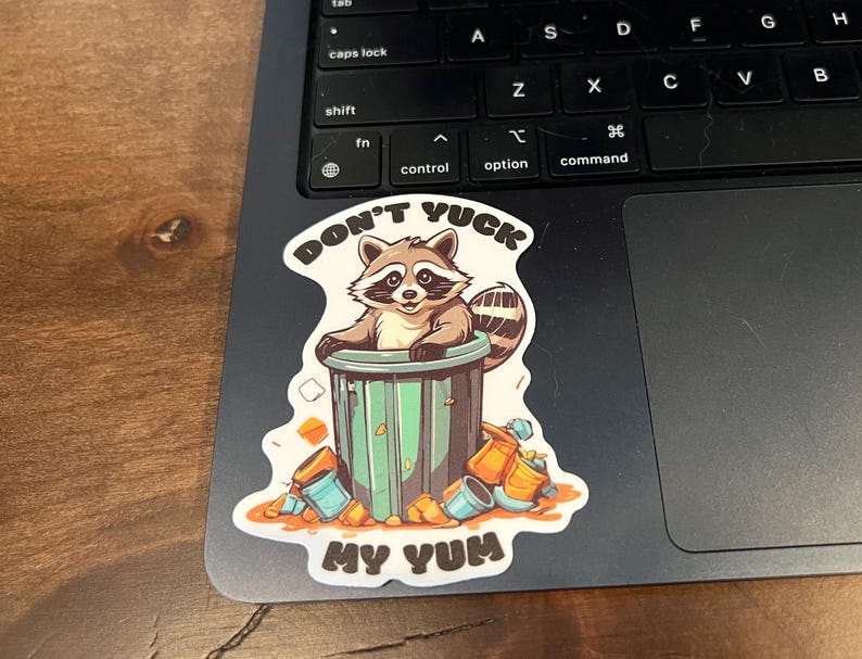 Sticker on black laptop. Sticker pictures cute raccoon in trash can with the words "Don't Yuck My Yum".