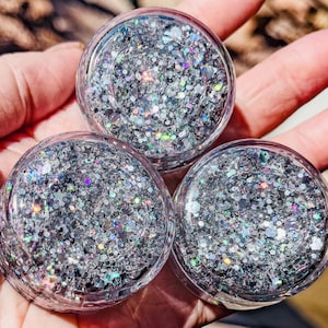 May include: Three clear, round containers filled with silver glitter. The glitter has a mix of shapes and sizes, including stars and iridescent pieces. The containers are held in a hand, with the glitter catching the light.