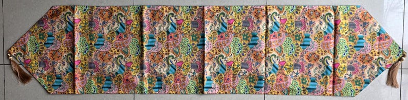 May include: A patterned table runner in shades of yellow, pink, blue, and green, featuring a paisley design. The runner has tassels at each end. The fabric is decorated with floral and abstract shapes.